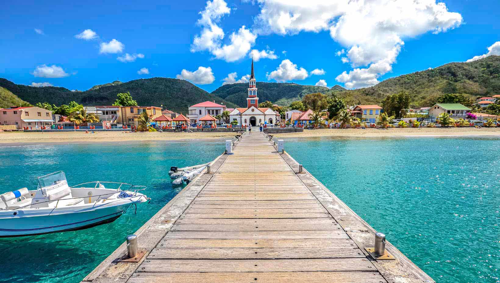 A wooden pier leading to a scenic coastal village with a colorful church and clear turquoise waters | MSC Cruises