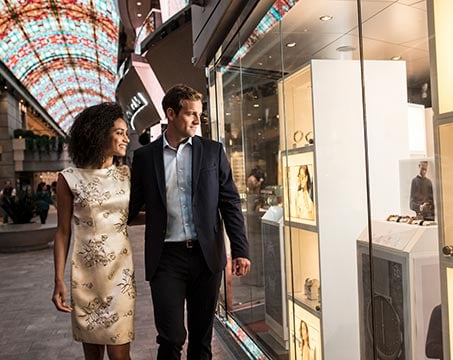 Couple strolling past luxury shops under a vibrant glass ceiling | MSC Cruises Couple strolling past luxury shops under a vibrant glass ceiling | MSC Cruises