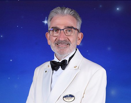 A crew member in a white formal uniform with an MSC badge against a starry blue background | MSC Cruises A crew member in a white formal uniform with an MSC badge against a starry blue background | MSC Cruises