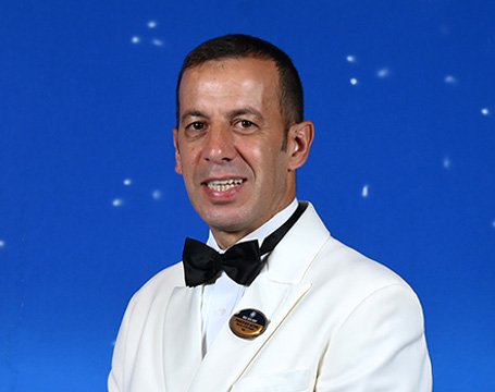 Crew member in a white uniform with a name badge against a starry blue background | MSC Cruises Crew member in a white uniform with a name badge against a starry blue background | MSC Cruises